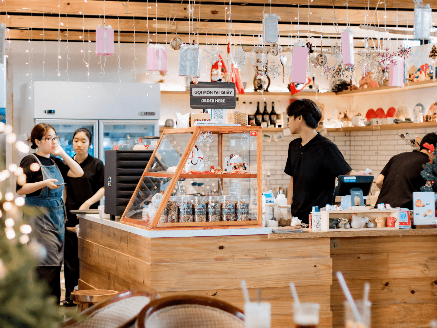 The interior of a warm, wood-paneled cafe bustling with staff. Several employees in black uniforms are behind a wooden counter featuring a glass display case filled with snacks. A sign above the counter reads "ORDER HERE" in both English and Vietnamese. The ceiling is festive, decorated with hanging string lights and pink gift-box ornaments.