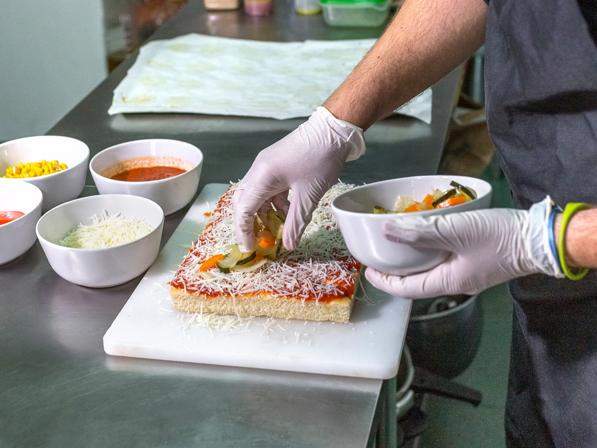 A close-up shot of a person wearing white gloves and a black apron preparing a rectangular pizza on a white cutting board. The pizza base is topped with tomato sauce and shredded cheese. The person is adding sliced vegetables from a white bowl. Several other bowls containing ingredients like corn and tomato sauce are visible on the stainless steel counter.