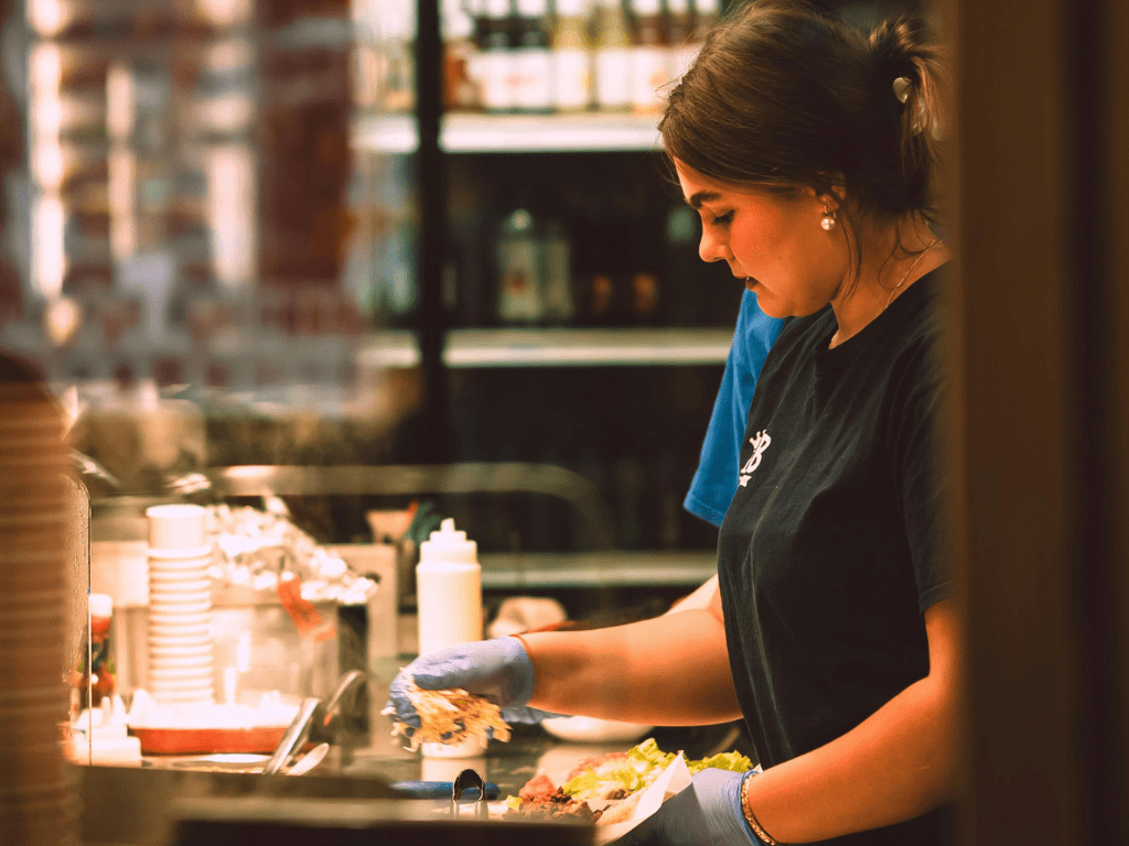 This image shows a food service worker in a busy kitchen or deli environment, focused on preparing a meal. The scene captures the fast-paced nature of the food industry while highlighting the importance of food safety and organization.