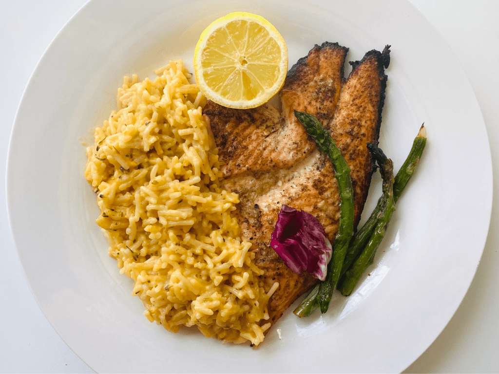 A top-down view of a white plate featuring seasoned grilled fish fillets, a side of yellow savory rice, grilled asparagus, and a fresh lemon slice.