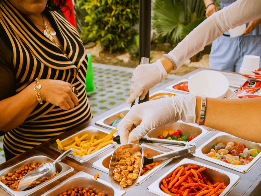 A close-up shot of a food service station outdoors where a person wearing white disposable gloves is using a metal scoop to serve snacks.