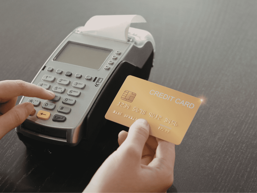 An over-the-shoulder view of a person completing a credit card transaction. One hand holds a gold-colored credit card with a visible chip and embossed numbers, while a finger on the other hand presses a button on a gray countertop payment terminal. A small receipt is beginning to print from the top of the machine.