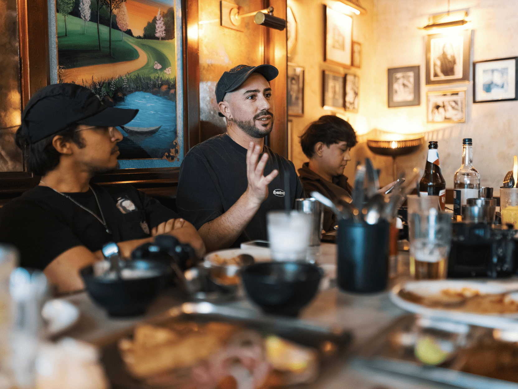 Group of people sitting around a table, engaged in discussion. One man gestures with his hands while speaking. Everyone wears black T-shirts and baseball caps. Plates of food and drinks are on the table.