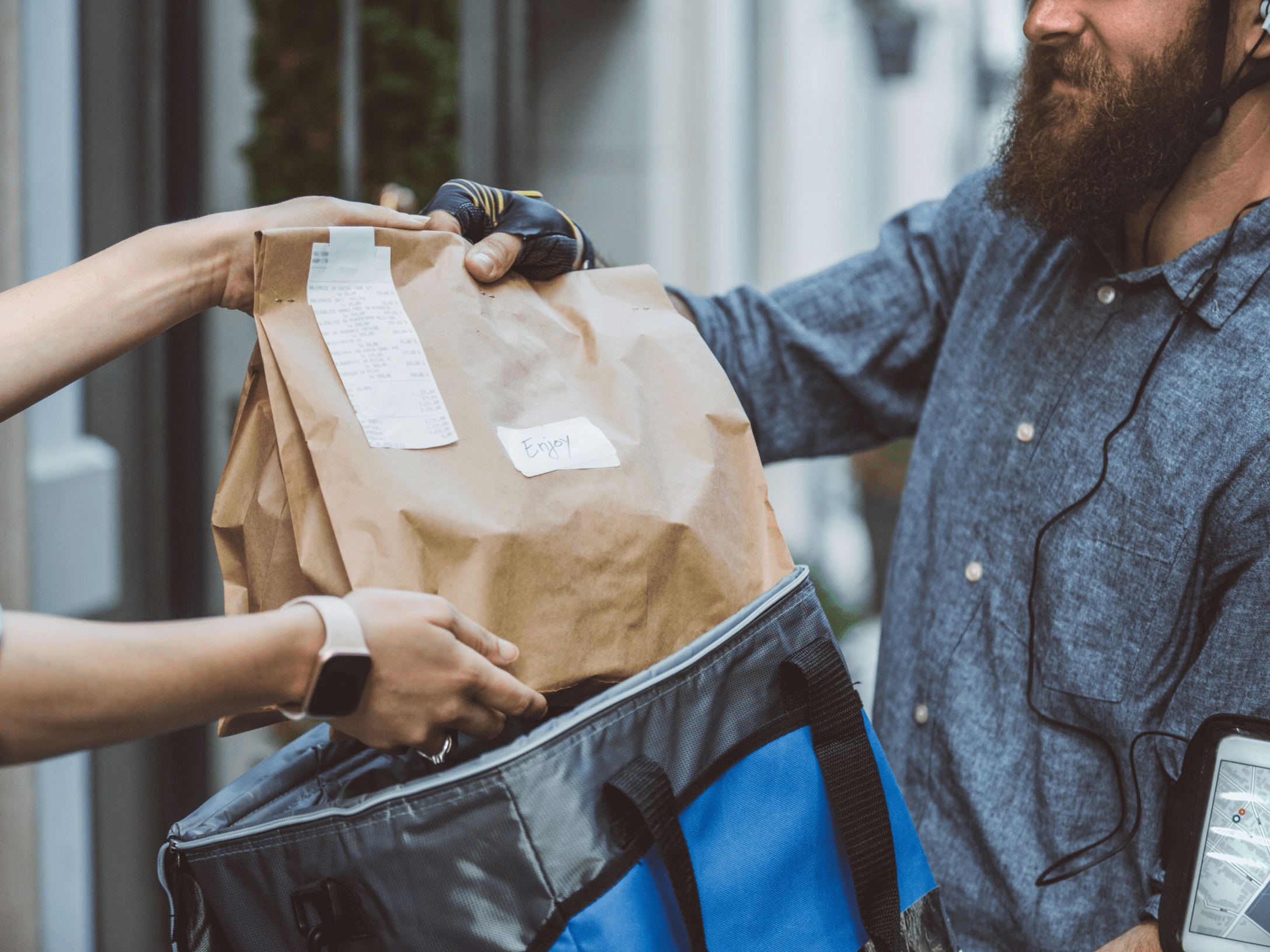 A close-up shot of a delivery person with a beard and a bike helmet handing a brown paper bag to a customer. The delivery person is wearing a grey shirt and black cycling gloves, and is pulling the bag out of a blue and grey insulated thermal bag. The customer, wearing a smartwatch, is reaching out with both hands to take the bag. The paper bag has a receipt stapled to it and a small white sticker that says "Enjoy." A smartphone displaying a map is visible on the delivery person's arm.